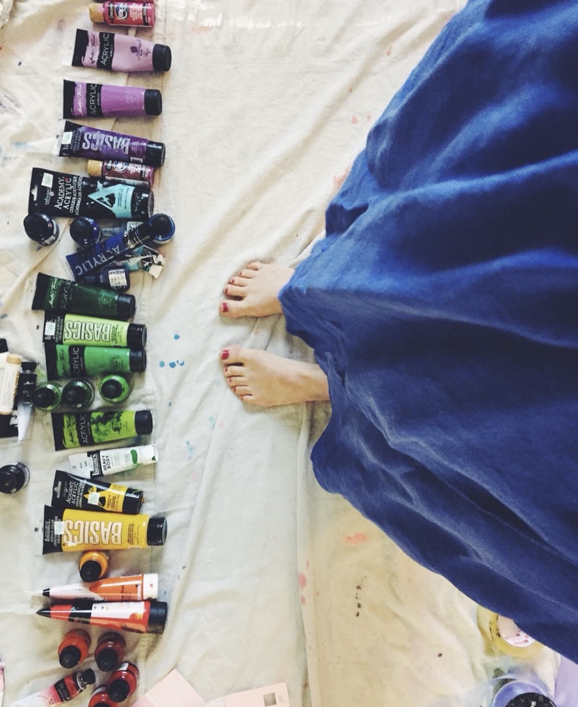 Bare feet with a red pedicure beside a row of paint tubes on a drop cloth, a person in a blue robe standing alongside