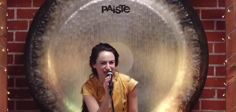 Alexandra Hulme seated before a large gong, chanting into a microphone